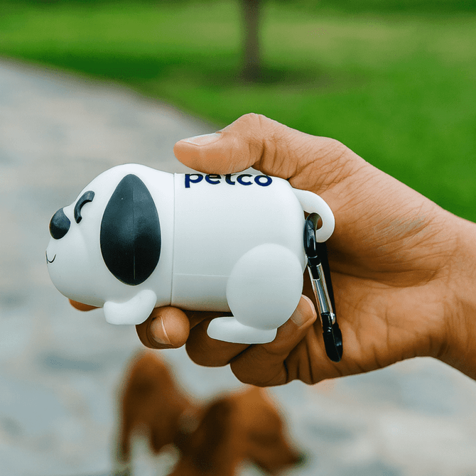 A white dog-shaped waste bag dispenser with black details, held in a hand, featuring a logo.