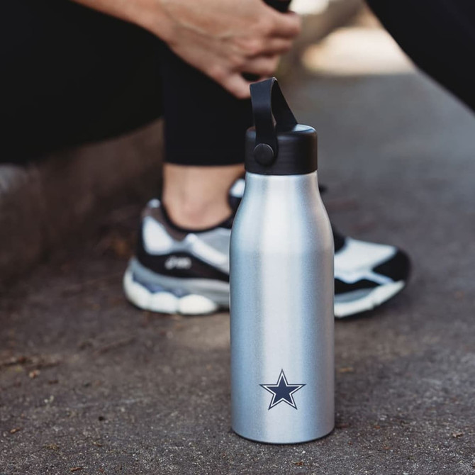 A drink bottle in silver with a logo, featuring a black lid and a sleek design, placed on a paved surface.