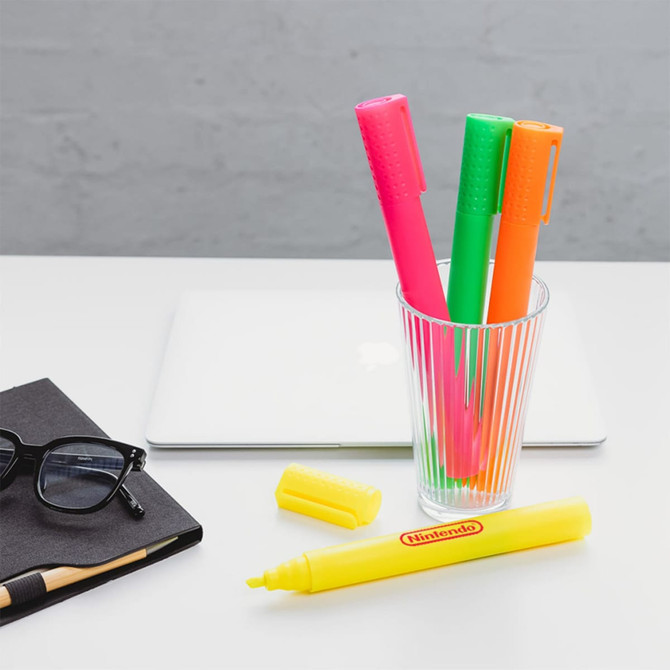 A glass holding pink, green, and orange jumbo highlighters alongside a yellow highlighter, with a notebook and glasses nearby. A glass holding pink, green, and orange jumbo highlighters alongside a yellow highlighter, with a notebook and glasses nearby.
