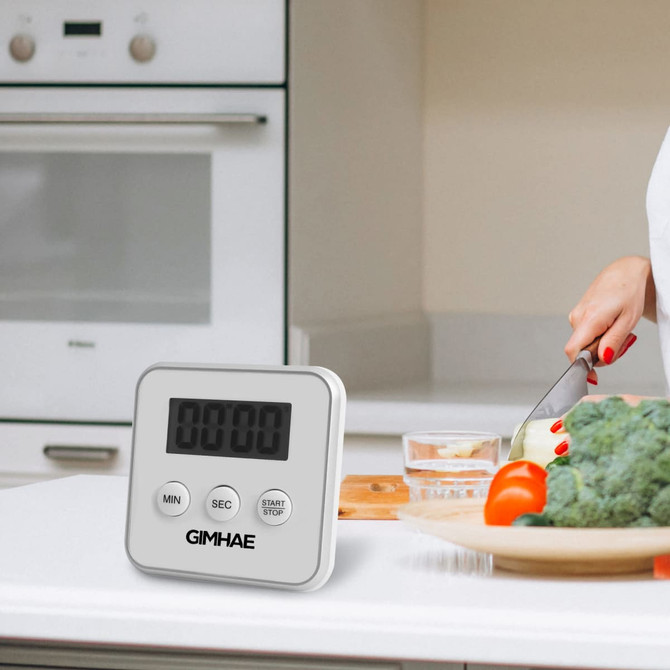 A digital kitchen timer in white with a large display, placed on a kitchen countertop beside fresh vegetables.