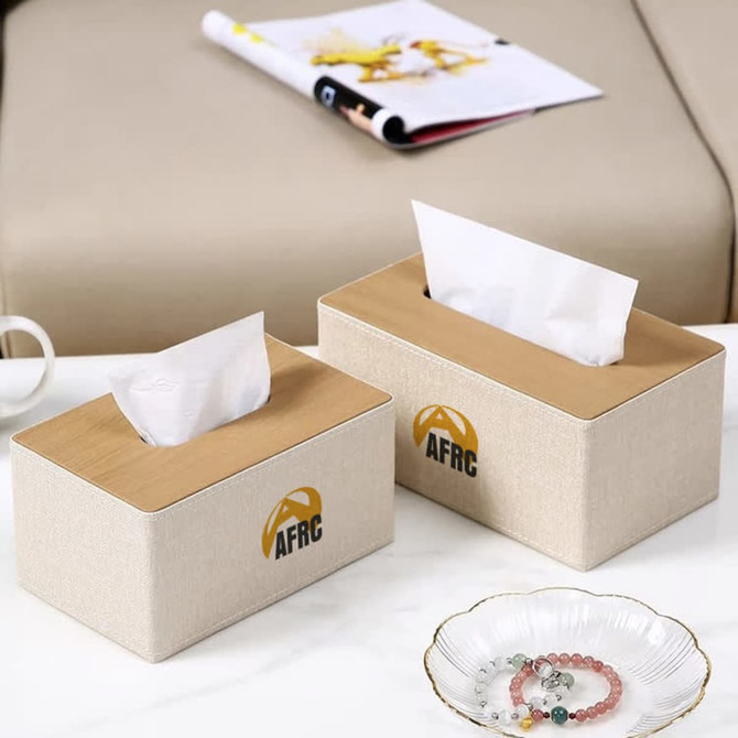 A medium tissue box set in beige with a wooden top and a logo, placed on a table alongside a magazine and a dish.