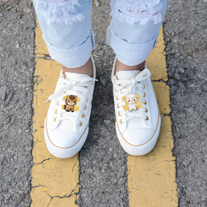 White shoes adorned with colourful charms featuring a bear and a cow, set against a grey pavement with yellow lines.