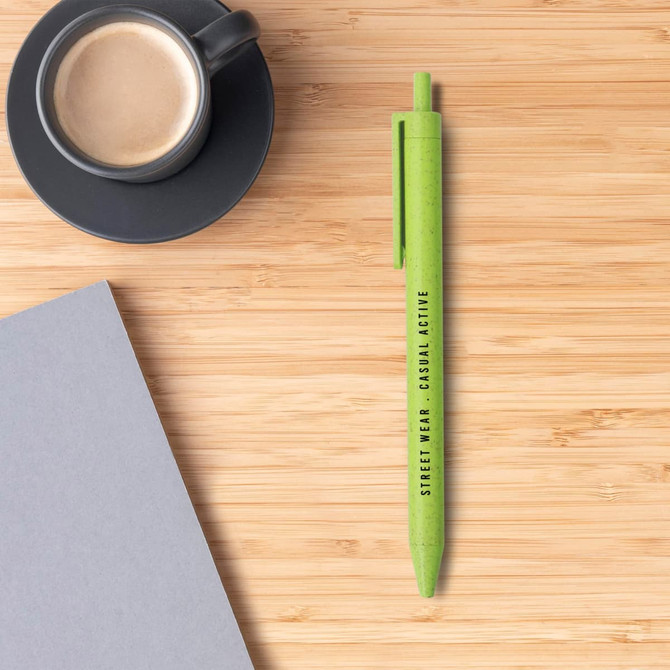 A green Wheat Straw ballpoint pen placed on a wooden surface next to a black coffee cup and grey paper.