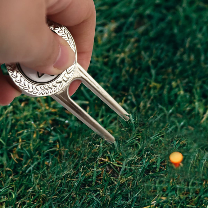 A silver divot repair tool with a ball marker is held above green grass, with a golf tee visible in the background.