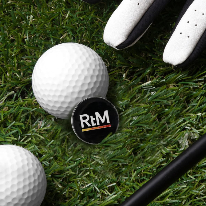 A round black golf ball marker situated on grass next to two white golf balls and a golf glove.