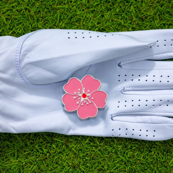A pink soft enamel golf ball marker featuring a floral design, placed on a white golf glove on green grass.