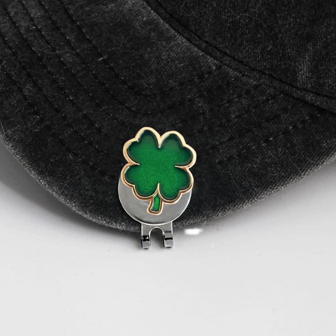 A green and gold soft enamel golf ball marker featuring a four-leaf clover design, resting on a black cap.