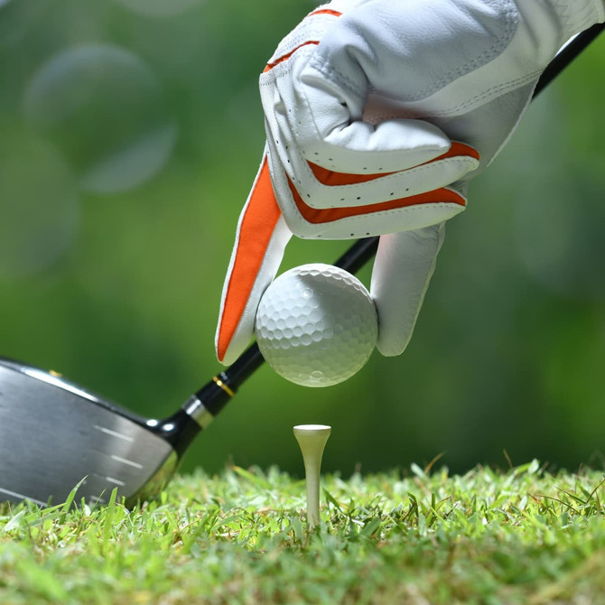 A hand in a white glove holds a golf ball above a bamboo golf tee on green grass, with a driver club in the background.