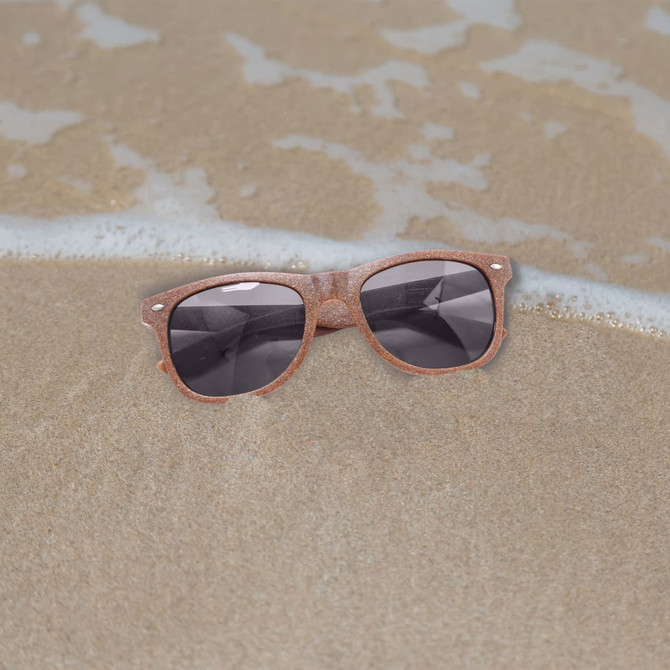 Coffee fibre sunglasses in a brown hue resting on sandy beach, with gentle waves in the background.