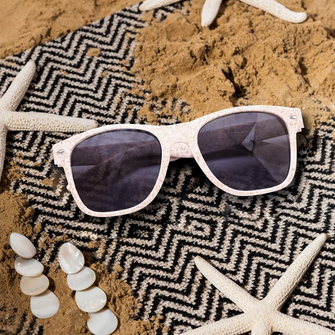 Wheat straw sunglasses in a light pink shade, resting on a sandy surface with starfish and seashells.