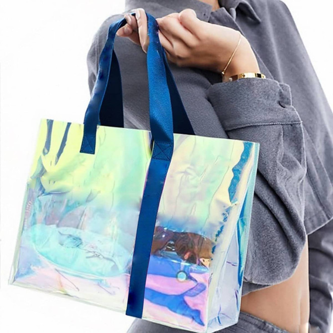 A transparent PVC tote bag with a colourful iridescent surface and navy blue handles, held by a person.