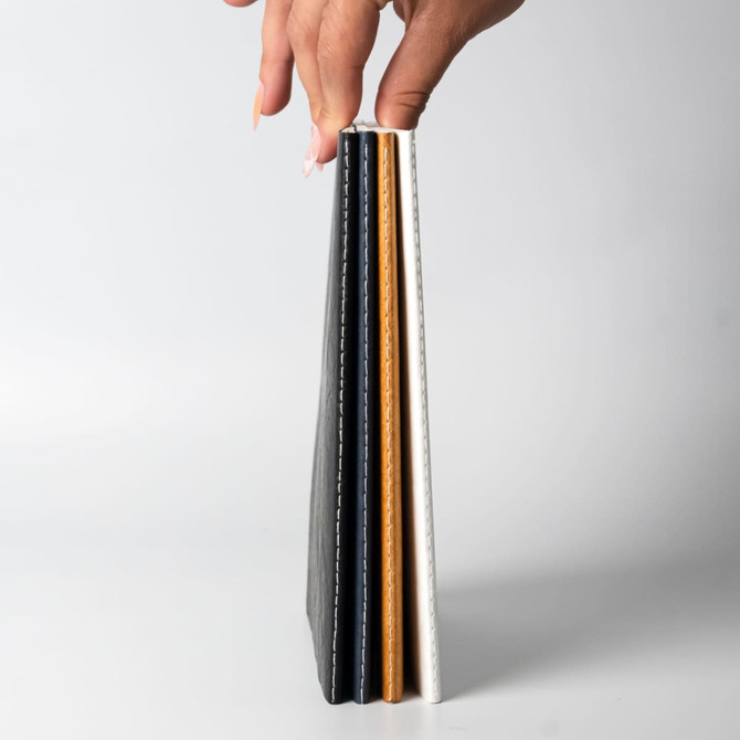 A hand holding a stack of three notebooks in black, tan, and white, featuring stitched edges.