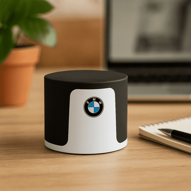 A black and white wireless speaker with a circular design and a logo, placed on a wooden desk near a plant and notebook. A black and white wireless speaker with a circular design and a logo, placed on a wooden desk near a plant and notebook.