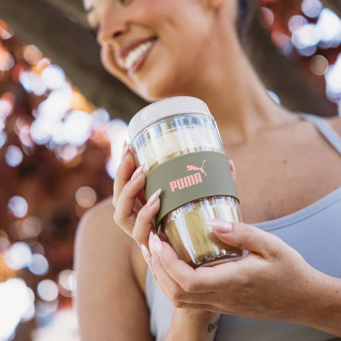 A reusable coffee cup with a transparent body and a green band, featuring a logo, held by a smiling person outdoors.