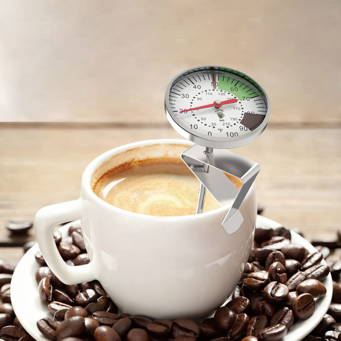 A silver instant read dial thermometer is placed in a steaming coffee cup, surrounded by coffee beans.