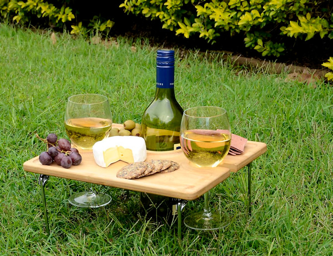A wooden picnic table with two glasses of white wine, grapes, olives, cheese, and crackers on a grassy surface.
