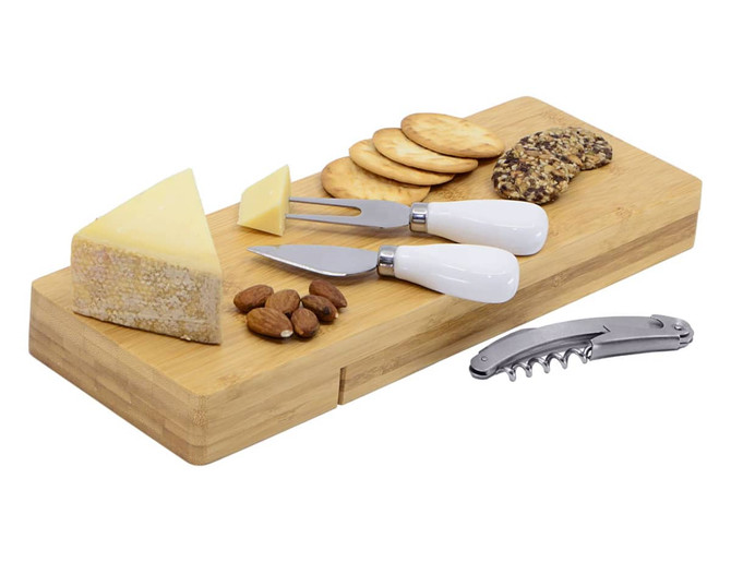 A bamboo cheese board with white-handled knives, various cheeses, nuts, crackers, and a corkscrew.