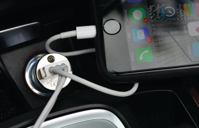 A phone charging cable connected to a silver car charger, with a smartphone displaying apps and notifications.