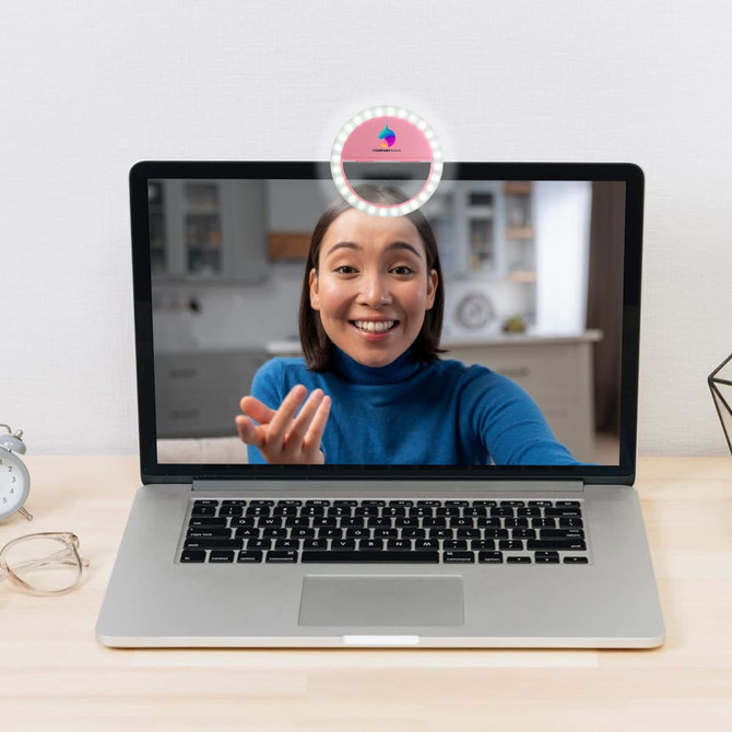 A ring selfie light on a laptop screen, illuminating a smiling woman in a blue top. The laptop is on a wooden desk.