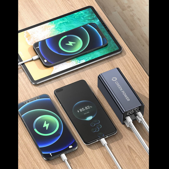 A black power bank with two cables, charging a tablet and two smartphones on a wooden surface.
