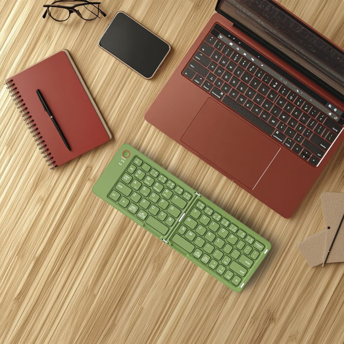 A green wireless foldable keyboard rests on a wooden surface next to a red laptop, notebook, pen, and phone.