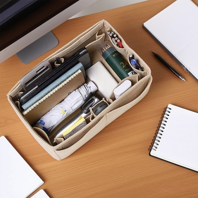 A beige felt bag organiser filled with notebooks, pens, a drink bottle, and a phone, set on a wooden desk.