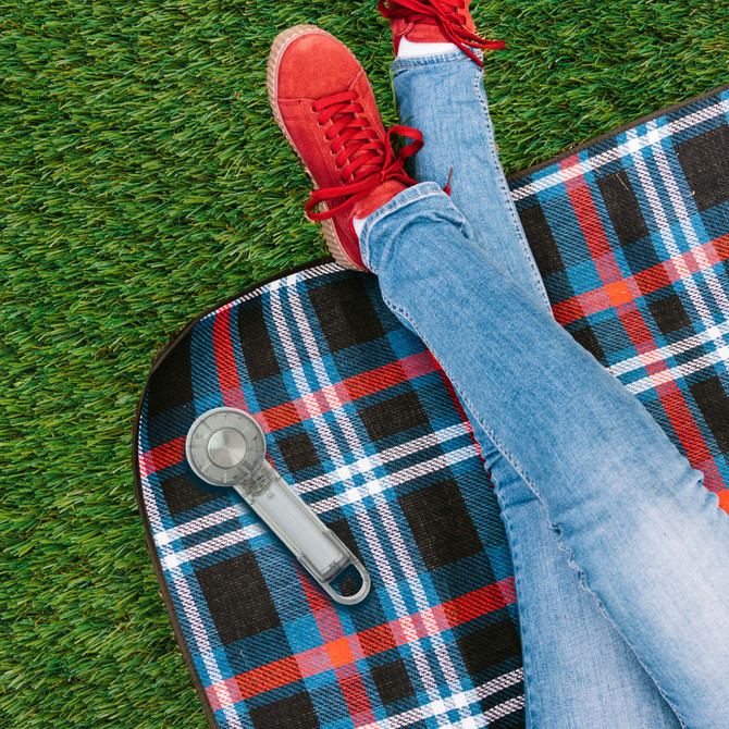 A transparent cooling fan sits on a checked picnic blanket beside a person wearing red sneakers and jeans.