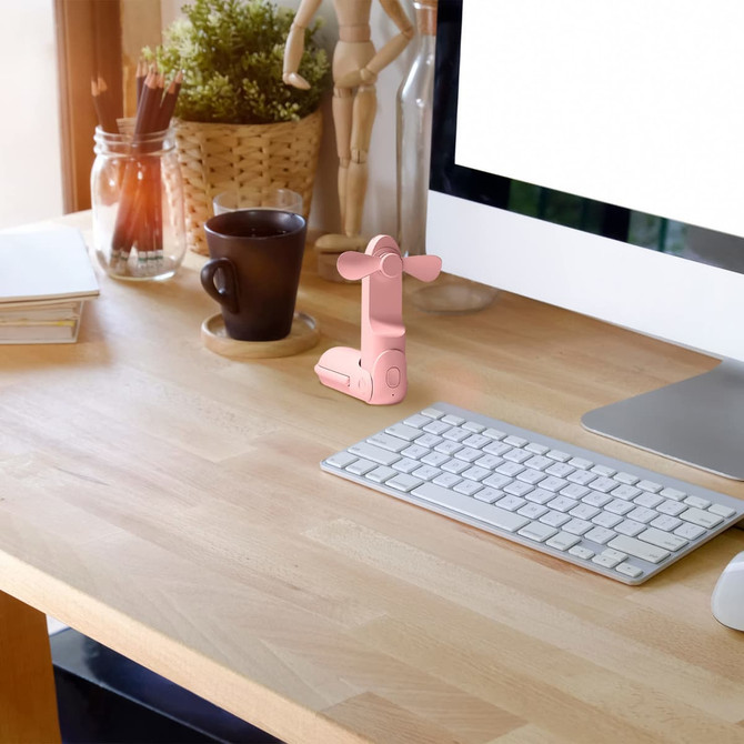 A pink handheld mini fan with a flashlight on a desk beside a coffee cup and a computer keyboard.