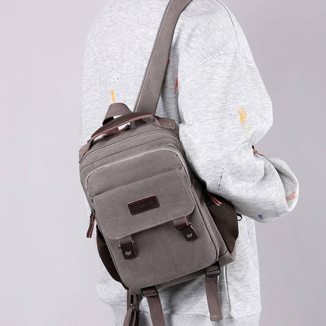 A grey canvas sling backpack hybrid with brown accents, worn on a person's shoulder. It has a logo on the front.