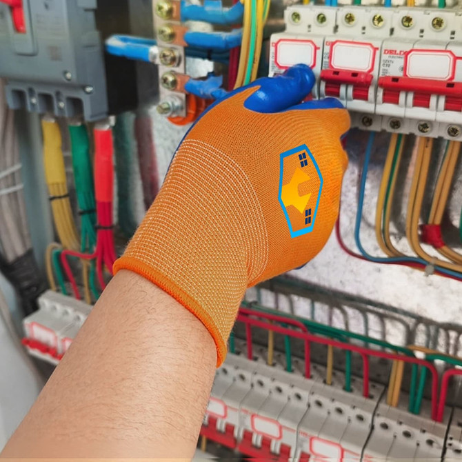 An orange insulating glove with a logo, placed on a hand near multicoloured electrical wiring and circuit breakers.