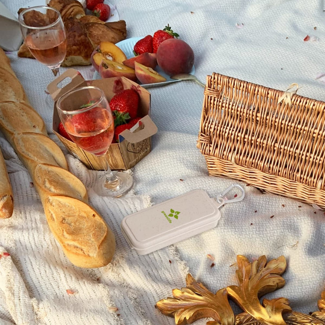 A white travel utensil set rests on a picnic blanket with a basket, glasses, bread, fruit, and pastries around it.