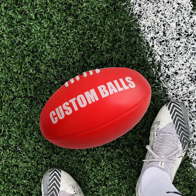 A red promotional AFL ball rests on artificial grass, beside a shoe with a sporty design. It features a logo.
