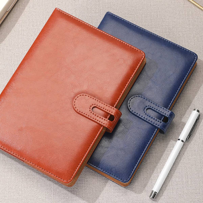 A brown and a navy blue PU notebook with a clasp, set on a textured surface beside a pen. Includes a logo.