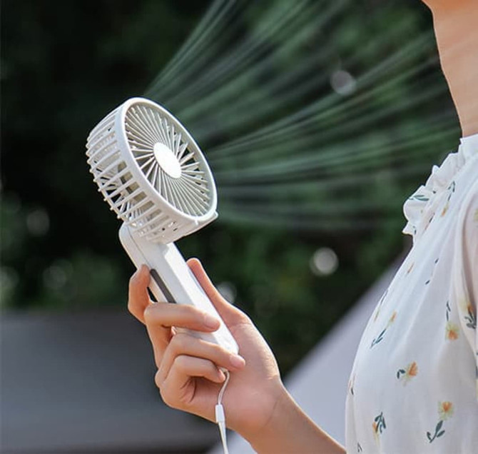 A white foldable USB hand fan is held in a person's hand, with air blowing from its grille. It features a logo.