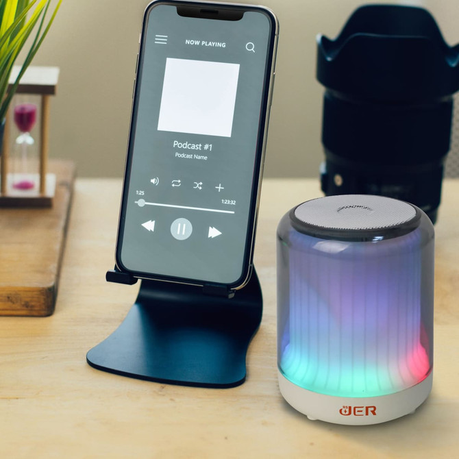 A portable Bluetooth speaker in multi-colours sits beside a smartphone on a stand, playing a podcast.