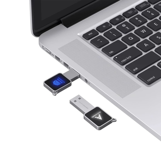 A square grey flash drive plugged into a laptop, featuring a logo and a blue light indicator.