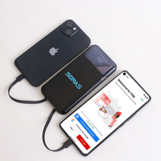 A black power bank with a logo, charging two smartphones on a light surface. The power bank has a sleek design.