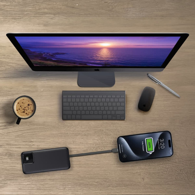 A black power bank with a charging cable connected to a smartphone on a desk with a computer, coffee, and a keyboard.