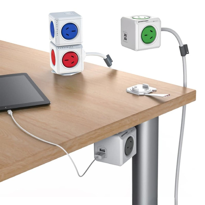 A Power Cube Extended Duo USB in white with red, blue, and green sockets, positioned on a wooden table alongside a tablet.