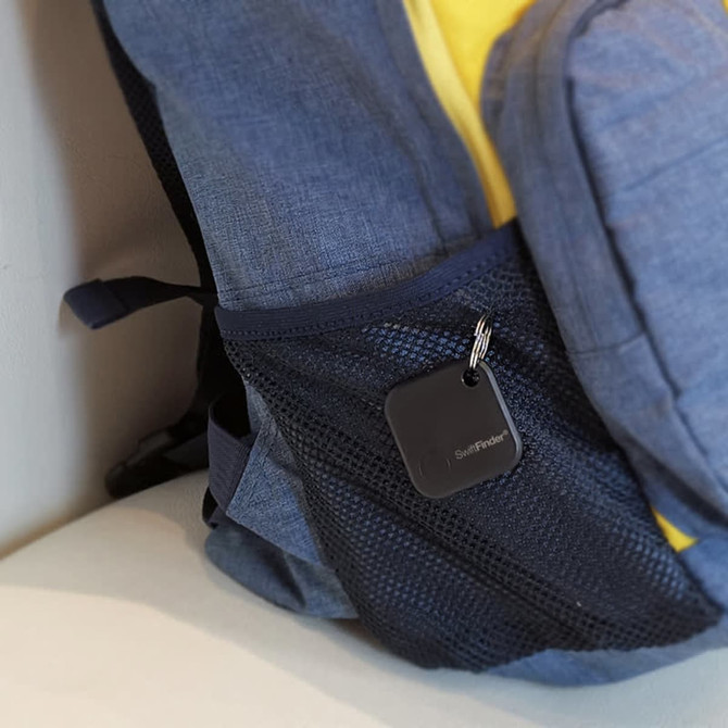 A black SwiftFinder Smart Tag Tracker attached to a dark mesh pocket of a blue and yellow backpack.