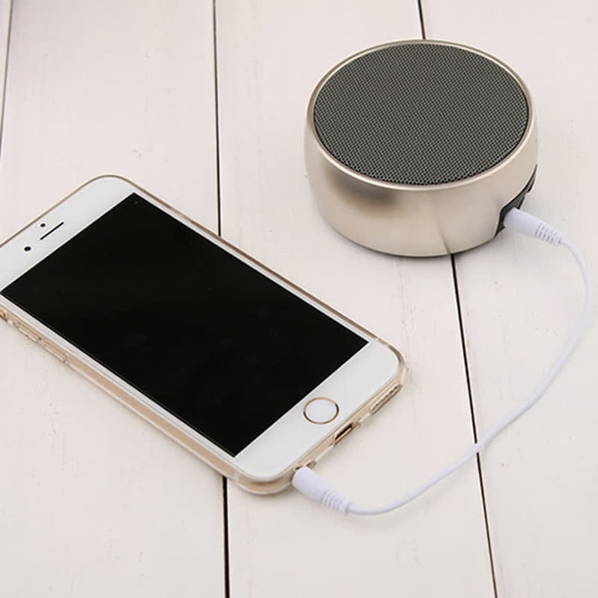A silver wireless speaker sits next to a smartphone, connected by a white audio cable on a wooden surface.