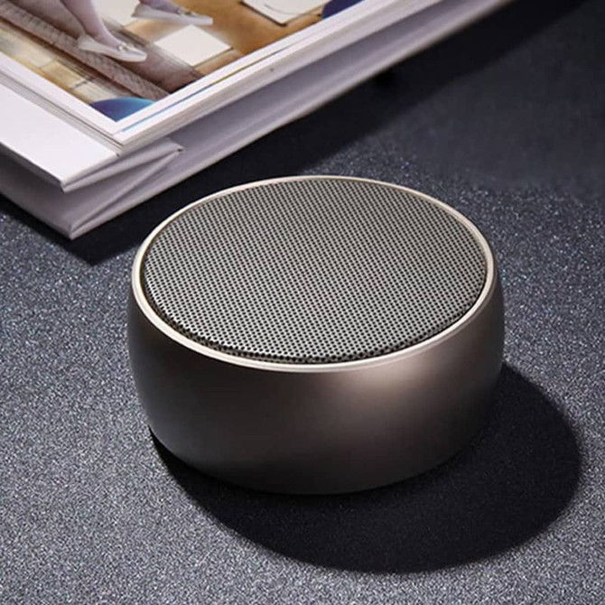 A round, grey wireless speaker with a textured grille, resting on a dark surface beside a stack of magazines.