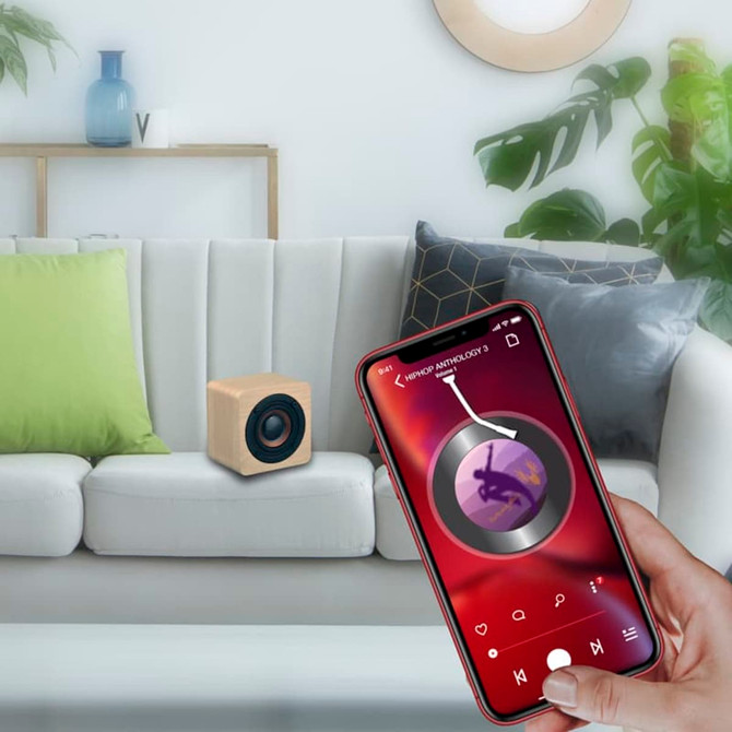 A wooden speaker sits on a white couch, with a smartphone displaying a music app in hand, featuring green and beige cushions.
