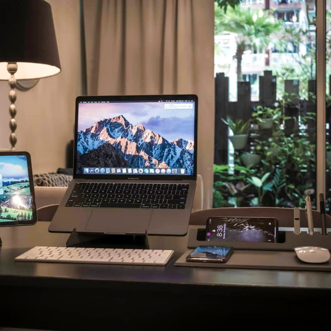 A sleek grey Ascend Mini Stand displays a laptop and phones, set on a stylish desk with greenery in the background.