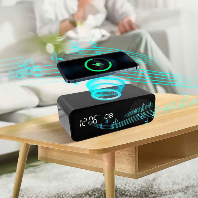 A wireless charging speaker in black, with a digital clock display, positioned on a wooden table beside a smartphone.