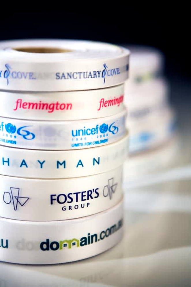 A stack of white ribbon rolls featuring various logos and branding in blue and black print. Each ribbon is 15mm wide.