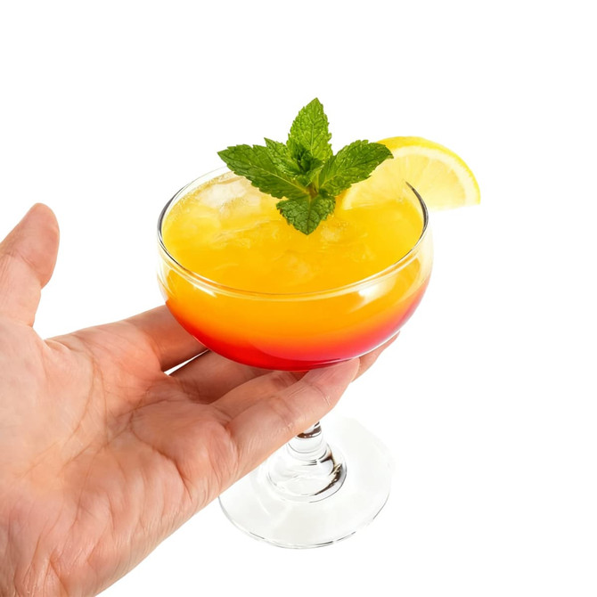 A hand holding a Martini coupe glass filled with a vibrant yellow and red drink, garnished with mint and a lemon slice.