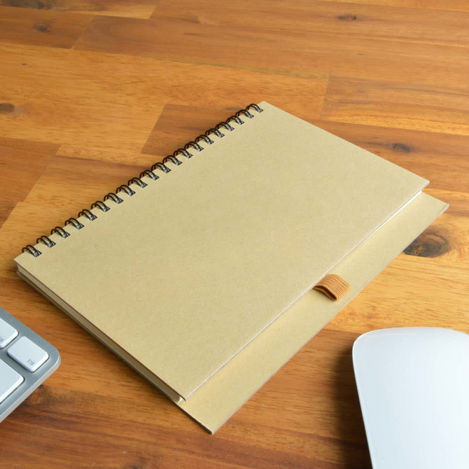 A spiral-bound notebook with a kraft cover, resting on a wooden surface next to a keyboard and mouse.
