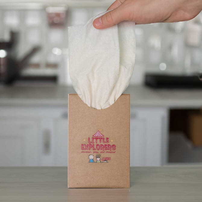 A hand holds a white tissue being pulled from a brown cardboard box with a playful logo.