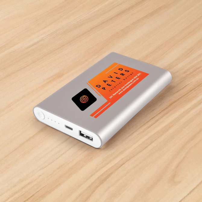 A silver power bank with a logo and an orange accent, featuring a USB port and indicator lights on a wooden surface.